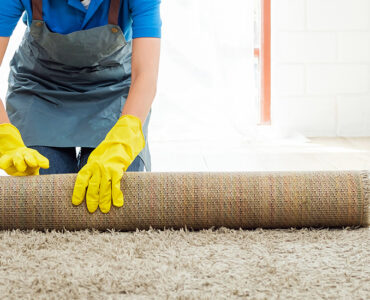 Smiling house cleaner wearing yellow gloves and apron rolling up a carpet in a bright, modern room. Professional home cleaning service concept for spotless, organized, and tidy living spaces.